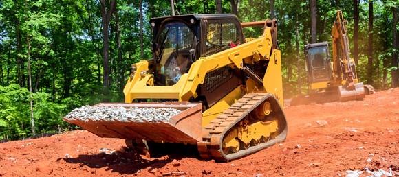 A track skid steer with gravel in it's bucket with a mini excavator in the background.